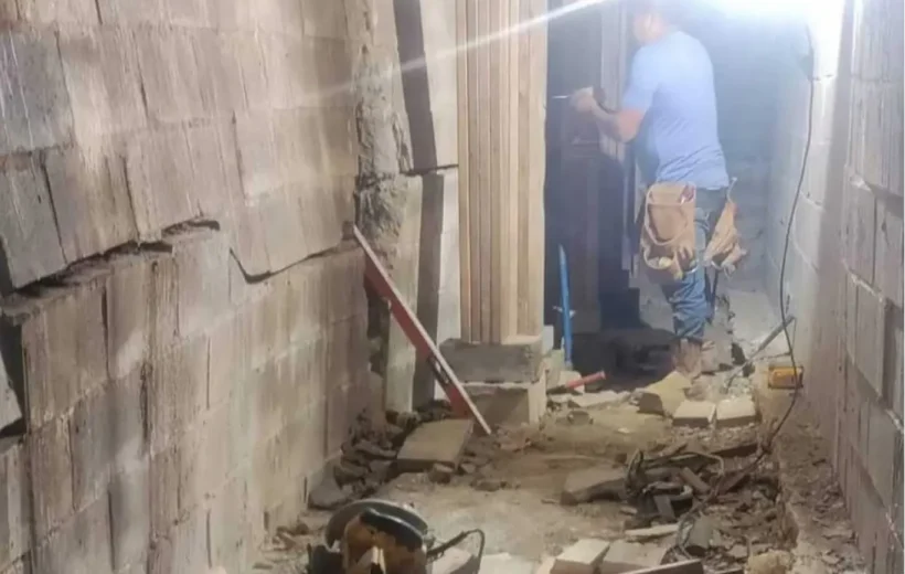 A construction worker in a blue t-shirt and tool belt works on a severely damaged cinder block basement wall in Louisville, KY. The left wall shows a significant horizontal crack and a noticeable inward bow, indicating structural failure. Debris and construction tools, including a circular saw and a level, are scattered on the floor amidst the ongoing repair work.