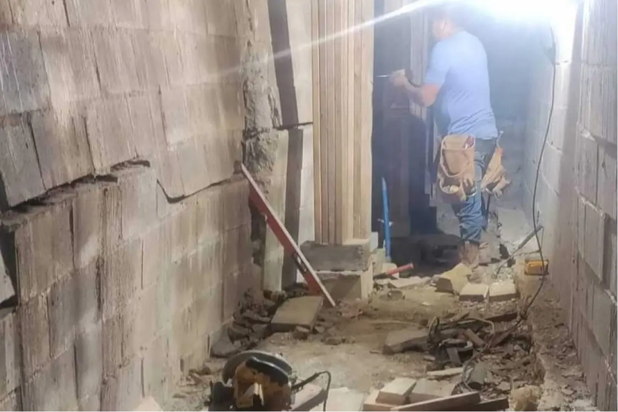 A construction worker in a blue t-shirt and tool belt works on a severely damaged cinder block basement wall in Louisville, KY. The left wall shows a significant horizontal crack and a noticeable inward bow, indicating structural failure. Debris and construction tools, including a circular saw and a level, are scattered on the floor amidst the ongoing repair work.