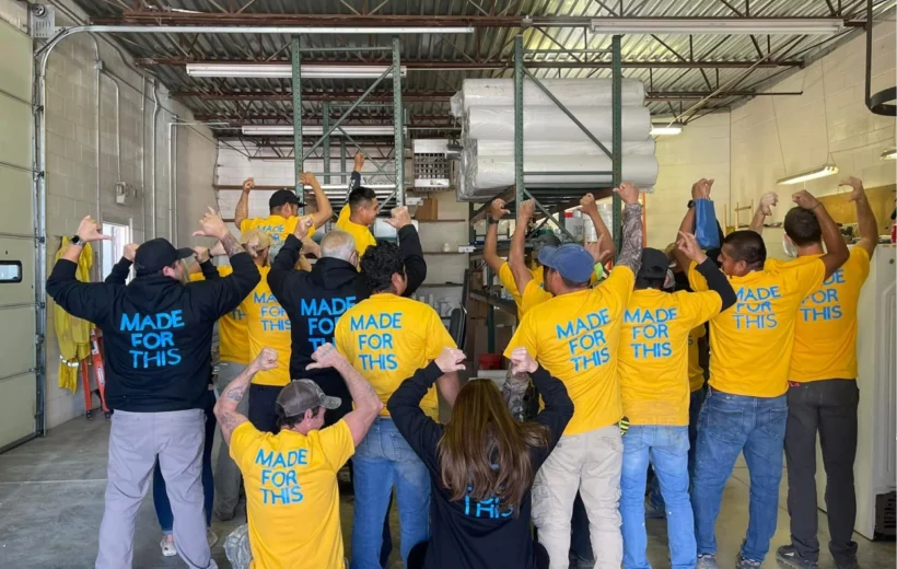 A large group of the Kentuckiana Contracting Services team stands in a warehouse, facing away from the camera to show off matching shirts that read "MADE FOR THIS" in bold blue letters.