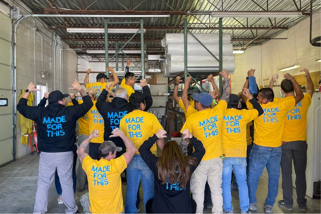 A large group of the Kentuckiana Contracting Services team stands in a warehouse, facing away from the camera to show off matching shirts that read "MADE FOR THIS" in bold blue letters.